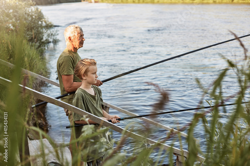 European grey haired mature father with son outdoors fishing by lake or river, standing near water with fishing rods in hands, dress casually, enjoying hobby and nature.