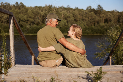 Father and son fishing together, sitting on wooden placement on bank of river, hugging and looking at each other, wearing casual attires, spending sunny day in open air.