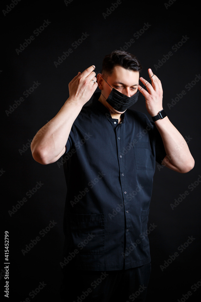 A young male doctor in a black surgical suit puts on a black mask to protect against the virus. Isolated on black background A young male doctor in a black surgical suit puts on a black mask to protect against the virus. Isolated on black background