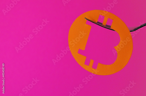 A bitcoin coin carved from orange paper hangs on a fork on a pink background with a copy space. Conceptual collage about cryptocurrency in the style of minimalism. High quality photo