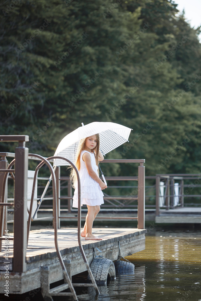 A beautiful girl of six years old in a short white dress stands on the masonry near the river and holds a white umbrella in her hands A beautiful girl of six years old in a short white dress stands on the masonry near the river and holds a white umbrella in her hands