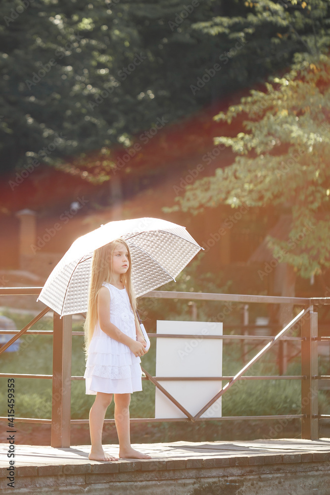 Fototapeta premium A beautiful girl of six years old in a short white dress stands on the masonry near the river and holds a white umbrella in her hands