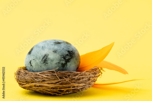 Easter blue egg and orange feathers in a nest on a yellow background. Easter holiday concept. Close up, copy space