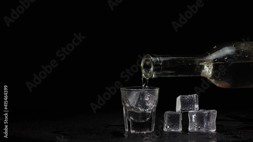 Shot glass on black table. Barman hand pours vodka, tequila or sake from bottle into empty shot glass on black background with ice cubes. Preparation to drink alcohol. Copy-space