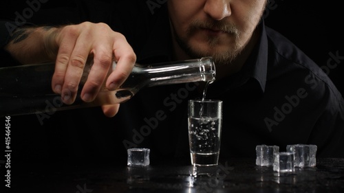 Alcoholic guy pouring up frozen vodka from bottle into shot glass with ice cubes on black background. Man drinking cold transparent alcohol drink sake, tequila or rum. Alcohol dependence addiction