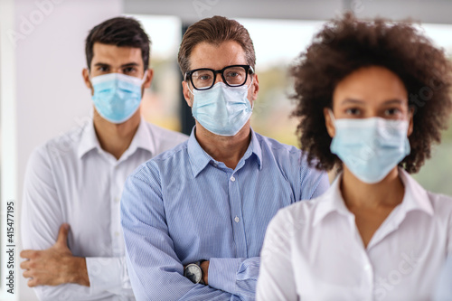 Multicultural group of business people with face masks standing with arms crossed in corporate firm during corona virus. Selective focus on man in the middle.
