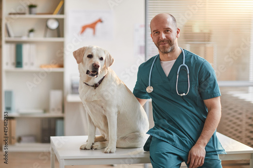 Portrait of mature male veterinarian smiling at camera while sitting on examination table with white Labrador dog at vet clinic, copy space