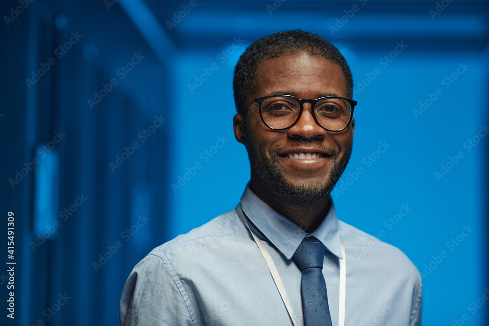 Portrait of smiling African American man wearing glasses and looking at camera while posing in server room at data center lit by blue light, copy space Portrait of smiling African American man wearing glasses and looking at camera while posing in server room at data center lit by blue light, copy space