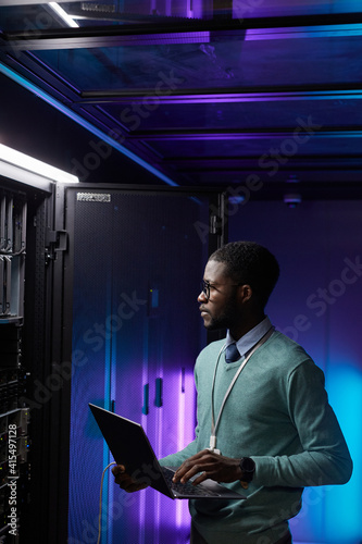Vertical portrait of young African American data engineer holding laptop while working with supercomputer in server room lit by blue light, copy space