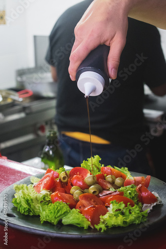 Una ensalada de lechuga, tomate y aceitunas mientras la mano de un hombre esparce una salsa sobre ella, servida en un plato de barro y de fondo se ven un fogón y otro hombre