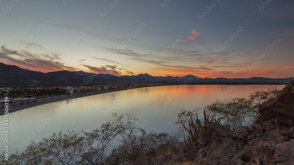 Sunset at the beach, orange sky and beachfront houses with mountains in the background and some clouds, soft reflections on the ocean, Loreto Bay, Mexico