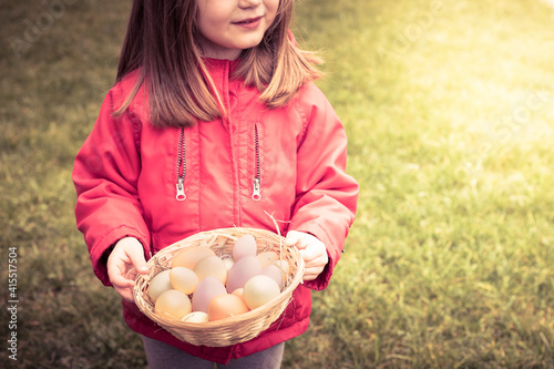 Little child girl  face not visible  wearing red jacket  smiling and holding a basket of eggs. Easter  spring wallpaper or background with sun flare and copy space.