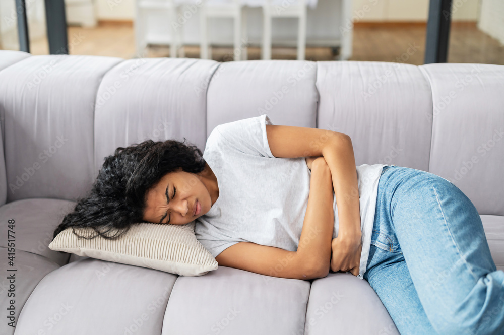 Young multiethnic woman with curly hairstyle is lying on a sofa and suffering from stomach ache, keeping hands on her stomach lying on the side, at home, waiting for doctor, poisoning, menstruation Young multiethnic woman with curly hairstyle is lying on a sofa and suffering from stomach ache, keeping hands on her stomach lying on the side, at home, waiting for doctor, poisoning, menstruation