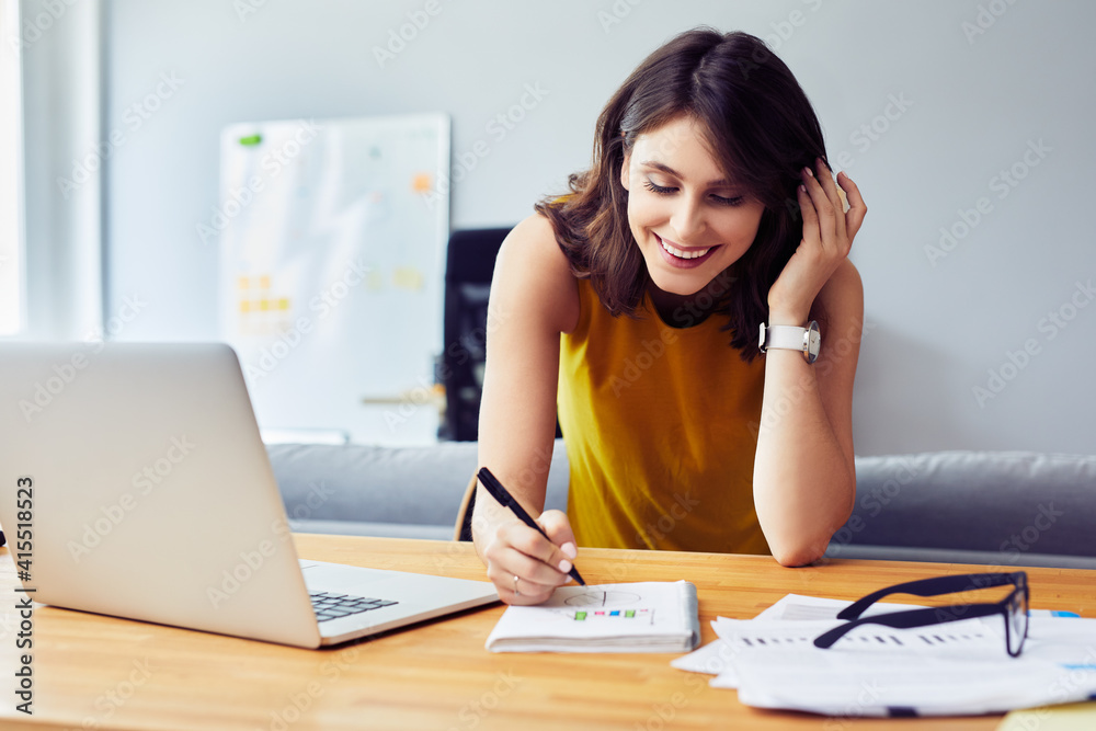 Beautiful smiling young entrepreneur making notes and using laptop in home office Beautiful smiling young entrepreneur making notes and using laptop in home office