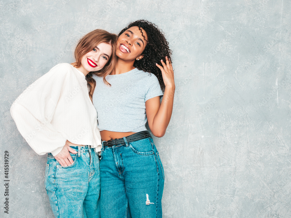 Two young beautiful smiling international hipster female in trendy summer jeans clothes. Sexy carefree women posing near gray wall in studio. Positive models having fun. Concept of friendship