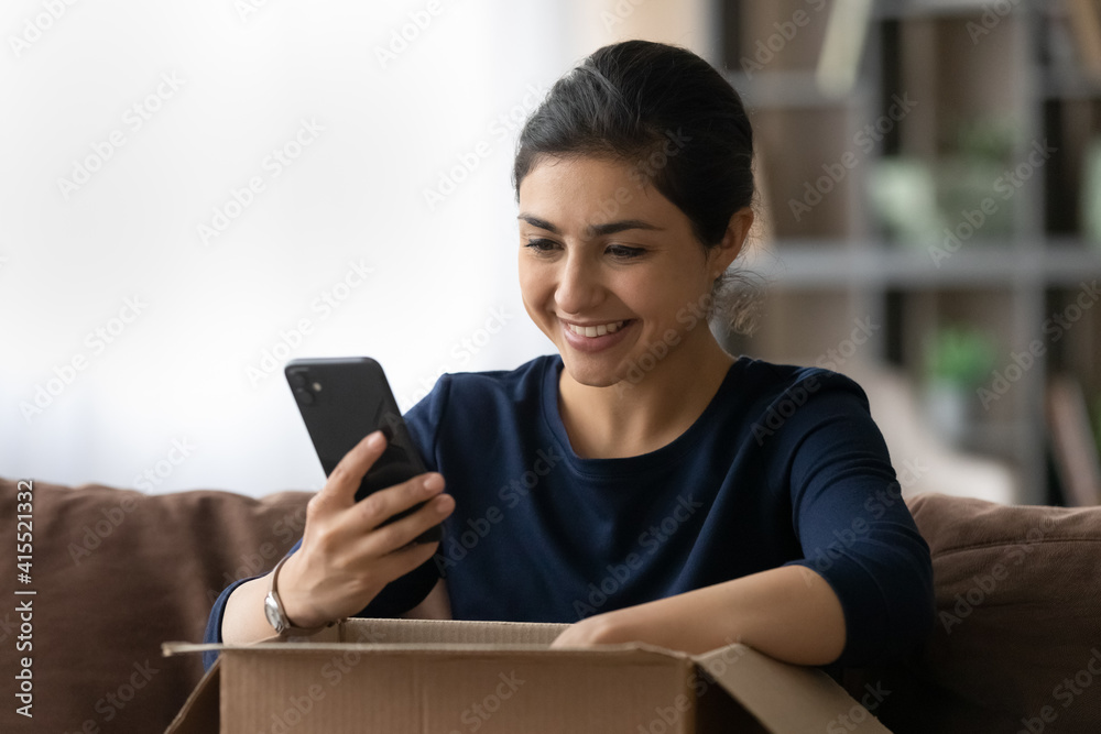 Happy young Indian woman unbox parcel with internet order, track or rate good delivery service on smartphone. Smiling ethnic female buyer client shop online on cellphone. Shipping concept. Happy young Indian woman unbox parcel with internet order, track or rate good delivery service on smartphone. Smiling ethnic female buyer client shop online on cellphone. Shipping concept.