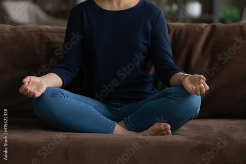 Crop close up of young Indian woman sit on sofa in living room with mudra hands meditate practice yoga at home. Mixed race ethnicity female relax rest relieve negative emotions. Peace concept.