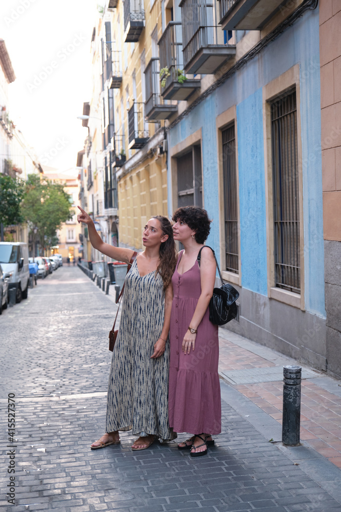 Beautiful lesbian couple wearing long dresses pointing and looking at something in the street. Young woman friends.