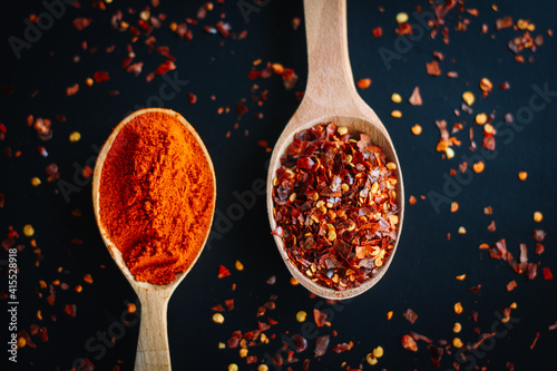 Fresh natural spices: red paprika and red chili pepper in wooden spoons on the black background. Top down view.