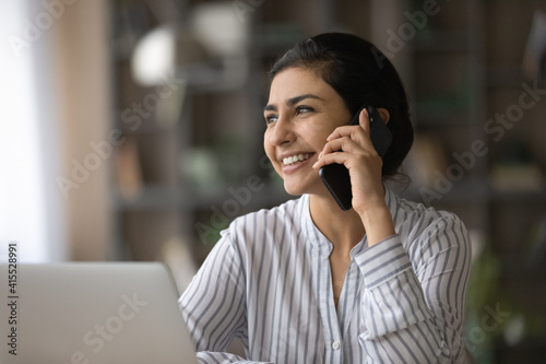 Smiling young Indian woman work on laptop talk speak on modern smartphone. Happy millennial mixed race ethnicity female have pleasant cellphone call, consult client online. Communication concept.
