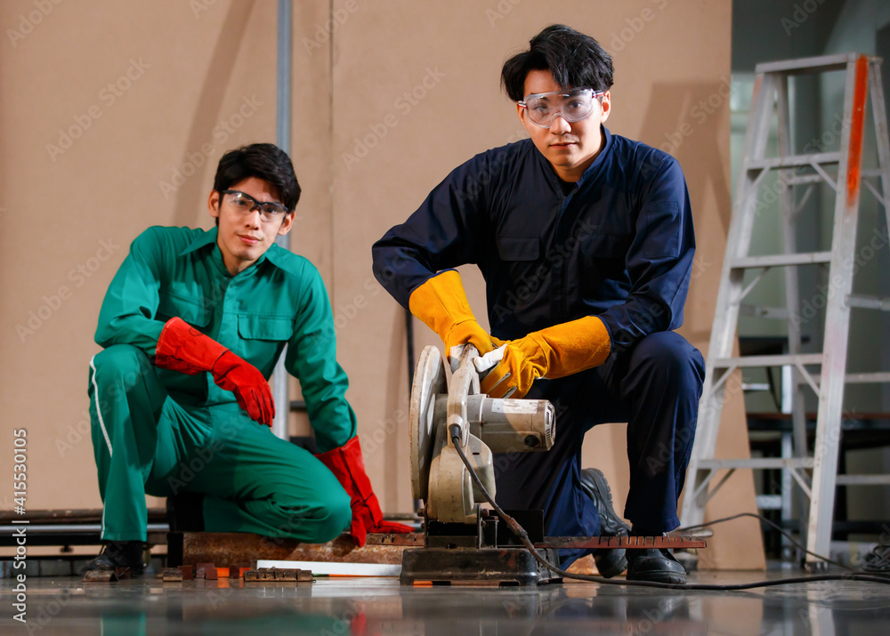 two young handsome Asian men wearing green and blue mechanic  jumpsuits, safety glasses, and gloves sitting in a factory workshop. Confident engineers mechanic working on a tool