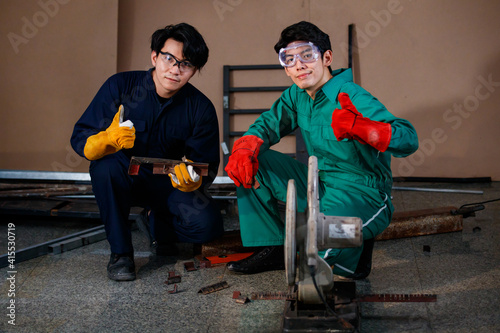 two smart Asian engineers wearing a mechanic jumpsuit, safety glasses, and glove sitting and holding tools posing smiling to a camera for photographing frow low angle view photo