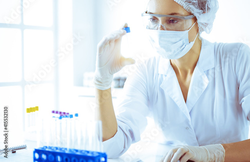 Professional female scientist in protective eyeglasses researching tube with reagents in sunny laboratory toned in blue. Medicine and science researching