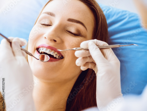 Smiling young woman with orthodontic brackets examined by dentist in sunny dental clinic. Healthy teeth and medicine concept