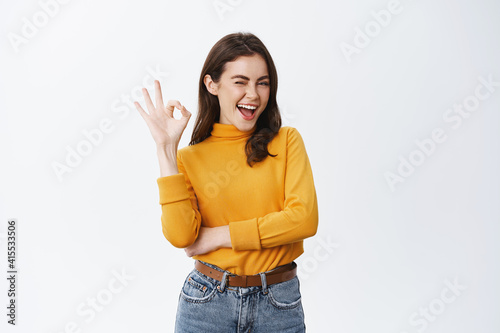 Confident young woman winking to assure you, showing okay sign and smiling, guarantee good quality, say alright or agree, like something or recommending, white background