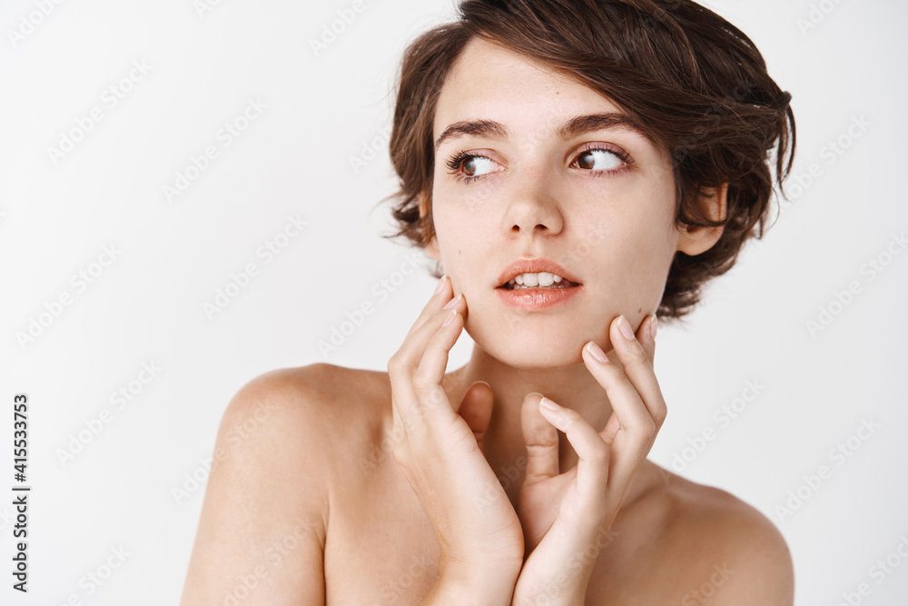 Close-up of tender young woman with naked shoulders, gently toughing natural no makeup facial skin, looking away pensive, standing over white background Close-up of tender young woman with naked shoulders, gently toughing natural no makeup facial skin, looking away pensive, standing over white background
