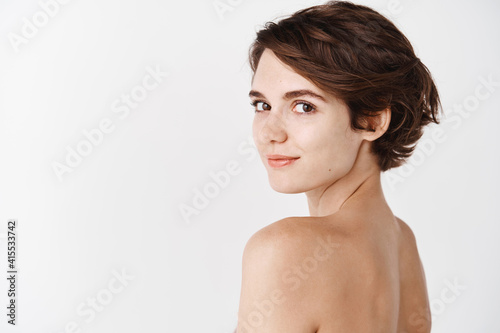 Skincare. Rear view of young caucasian woman turn head back at camera, standing half naked on white background and smiling. Tender girl with no makeup and natural beauty