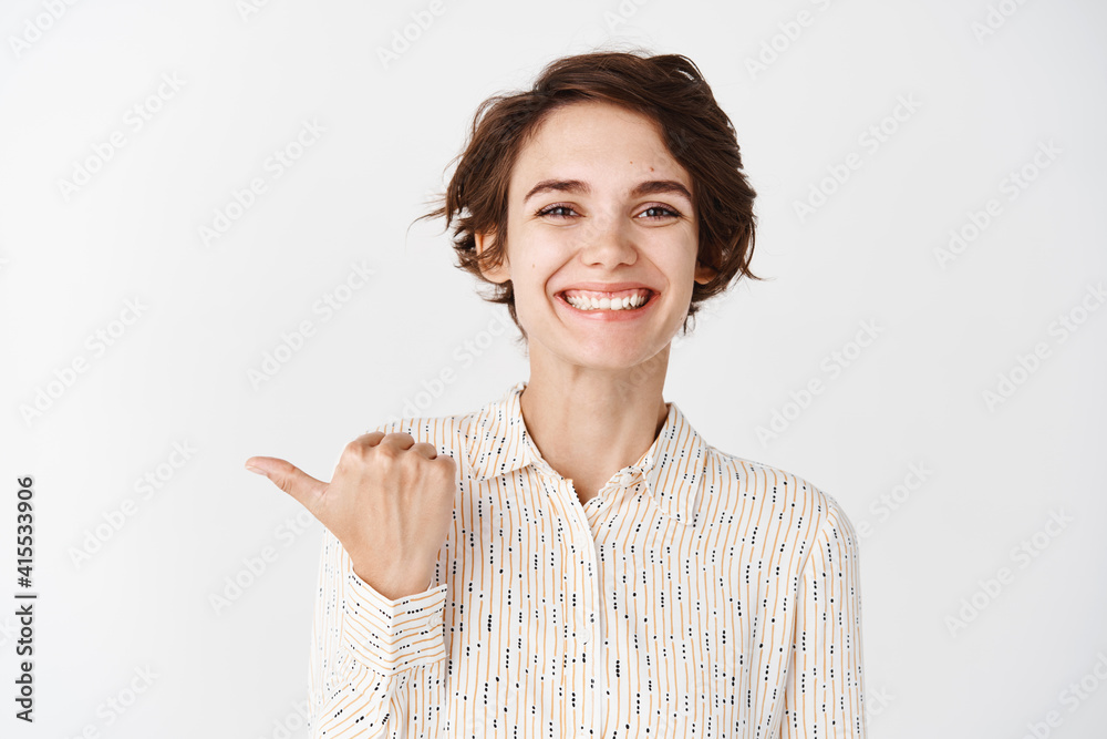 Young smiling woman employee looking happy and proud, pointing finger left at logo company, standing against white background Young smiling woman employee looking happy and proud, pointing finger left at logo company, standing against white background