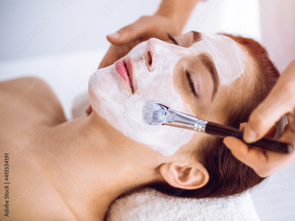 Beautiful brunette woman enjoying applying cosmetic mask with closed eyes in sunny spa center. Relaxing treatment and medicine concept
