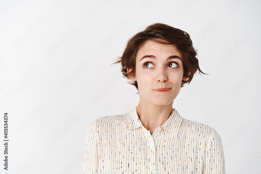 Cute young woman with shirt haircut, looking tempted at upper left corner empty space, thinking about something, standing pensive on white background Cute young woman with shirt haircut, looking tempted at upper left corner empty space, thinking about something, standing pensive on white background