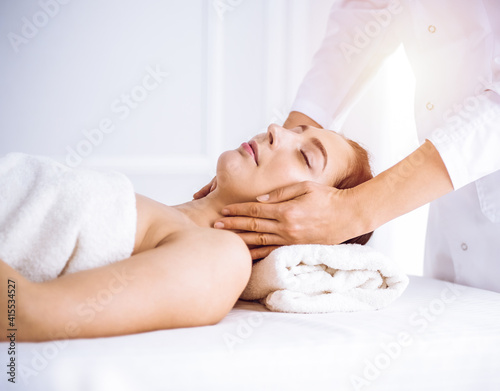 Beautiful brunette woman enjoying facial massage with closed eyes in sunny spa center . Relaxing treatment and cosmetic medicine concepts