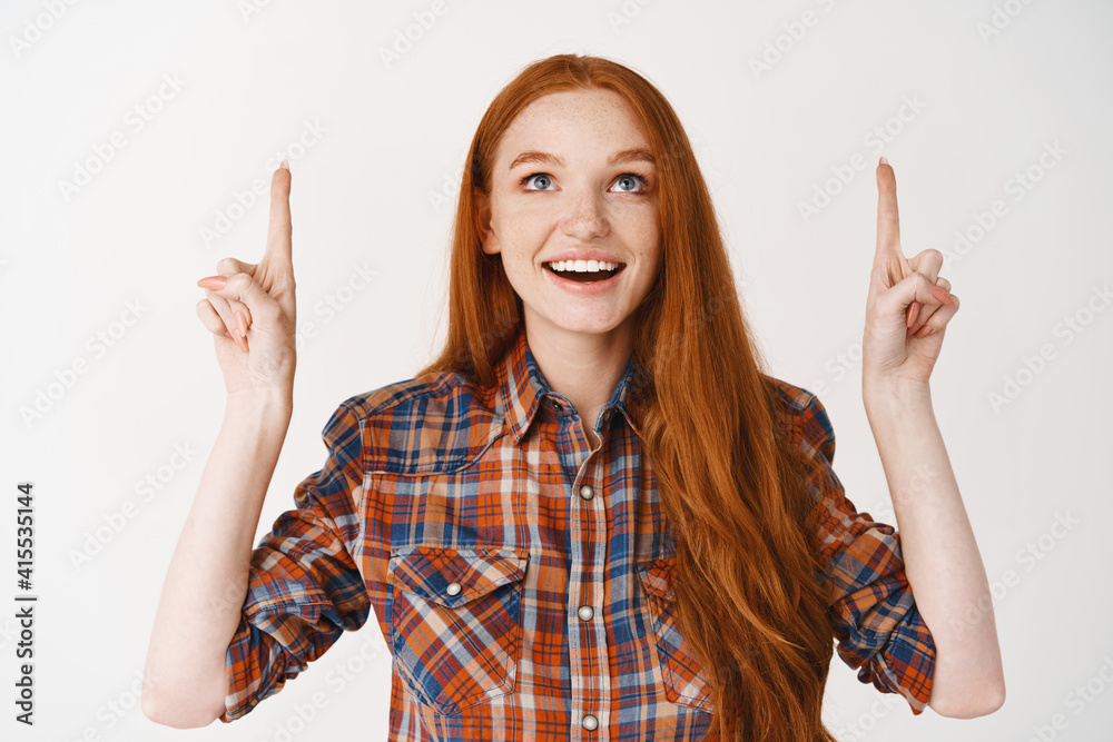 Beautiful teenage girl with red long hair, looking and pointing fingers up amazed, standing over white background