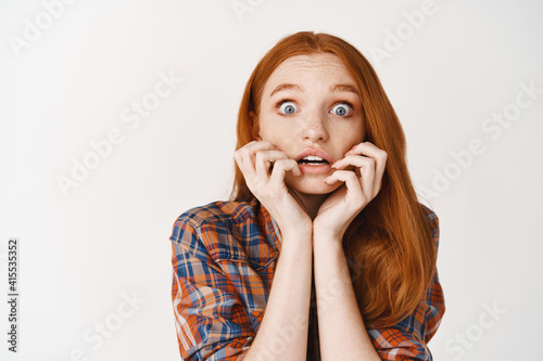 Close-up of amazed redhead girl listening with interesting, biting fingernails tempted and amused, looking at camera, standing over white background