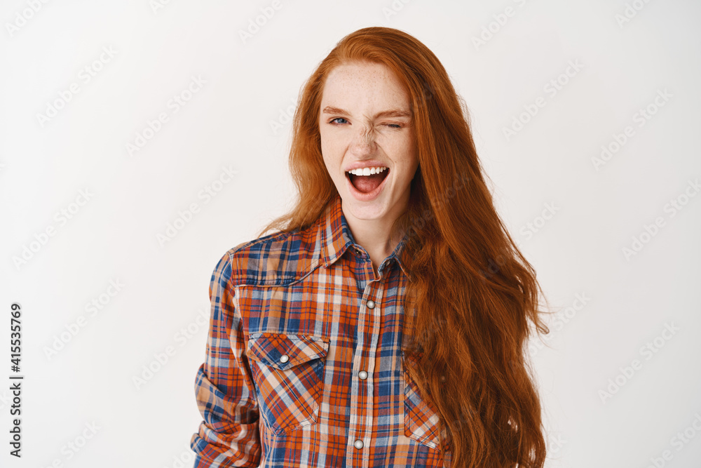 Attractive girl with ginger hair and blue eyes winking at camera assuringly, standing over white background Attractive girl with ginger hair and blue eyes winking at camera assuringly, standing over white background