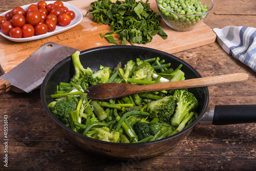 Cooking green vegetables for pasta in a frying pan - beans, broccoli, asparagus in a frying pan on a wooden table, tomatoes and chopped spinach on a board