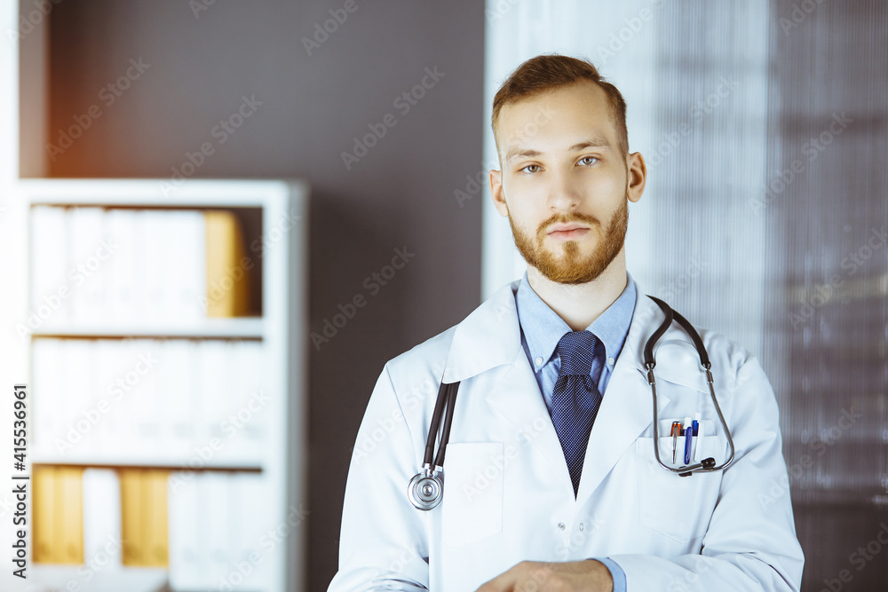 Friendly red-bearded doctor using tablet computer in sunny clinic at his working place. Medicine concept