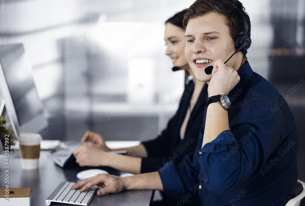 Two young people in headsets are talking to the clients, while sitting at the desk in a modern office. Focus on man in a blue shirt. Call center operators at work Two young people in headsets are talking to the clients, while sitting at the desk in a modern office. Focus on man in a blue shirt. Call center operators at work