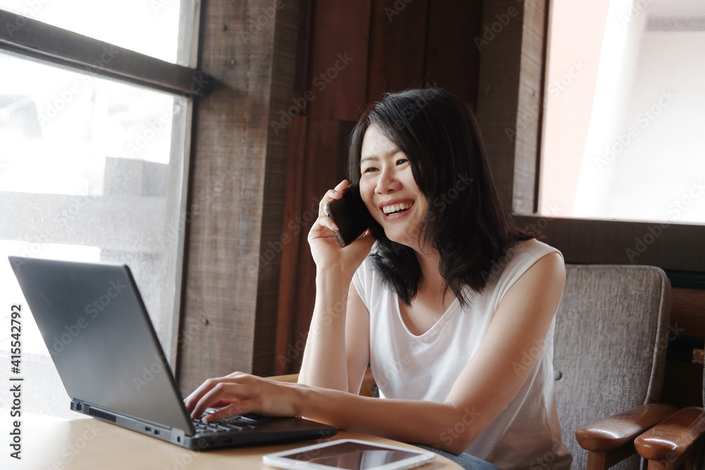 Smiling young Asian woman is calling with smartphone and online working with laptop in living room. Work from home for Covod-19 outbreak concept. Smiling young Asian woman is calling with smartphone and online working with laptop in living room. Work from home for Covod-19 outbreak concept.