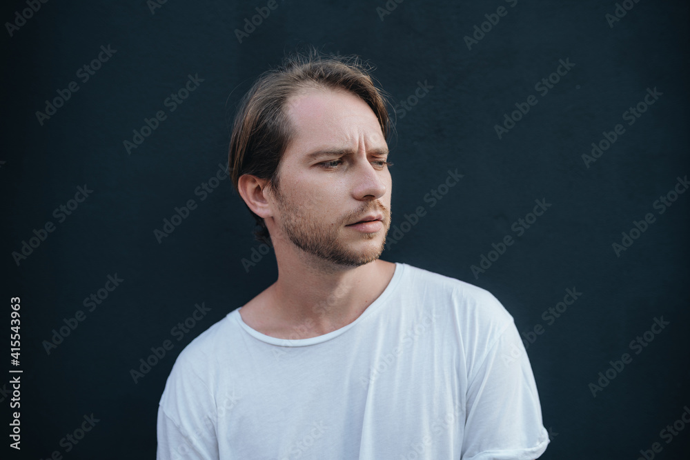 Portrait of frowned european man in white t-shirt at dark wall