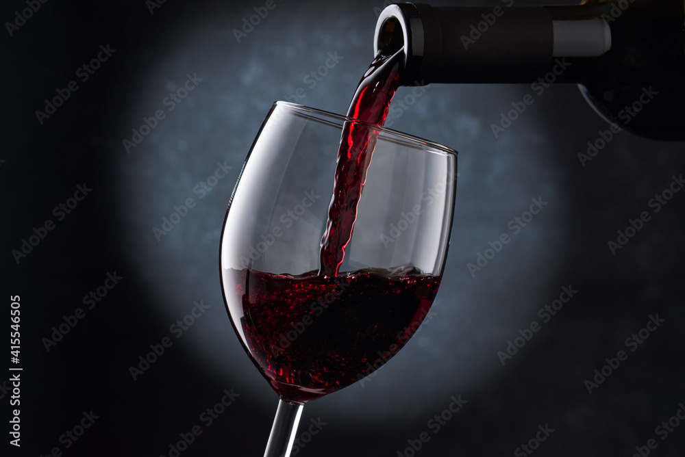 Fototapeta premium Red wine is poured into a glass from a bottle on a blurry blue background, a stream of red wine from the bottle swirls in the glass, close-up. Free space for text.
