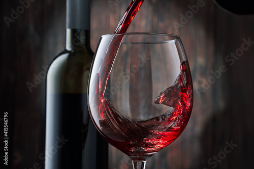 Red wine is poured into a glass from a bottle on a blurred wooden background, a stream of red wine from the bottle swirls in the glass, close-up. Free space for text.