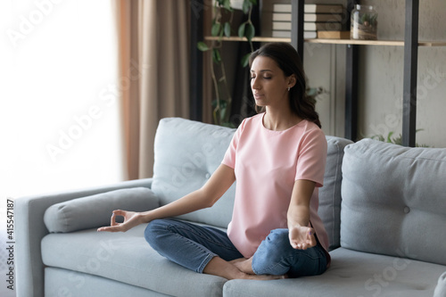 Concentrated young indian female sitting on sofa with crossed legs practicing yoga pranayama exercises. Calm relaxed millennial arabic woman meditating at home taking lotus position relieving stress