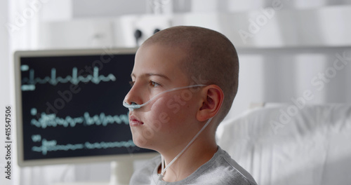 Konstfotografi Side view of teen boy with cancer sitting in bed with vital signs on computer mo