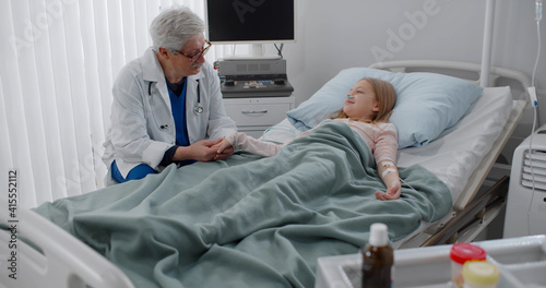 Senior male doctor holding hand and talking to sick girl patient resting in hospital bed