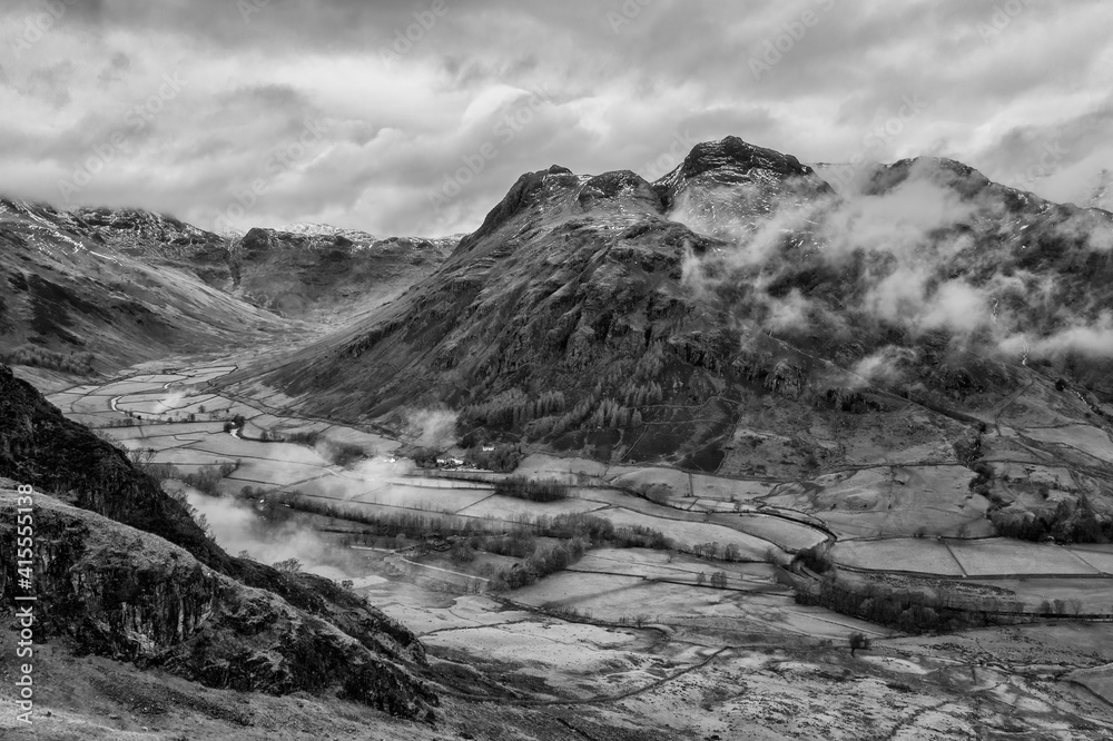 Stunning flying drone  black and white landscape image of Langdale pikes and valley in Winter with dramatic low level clouds and mist swirling around