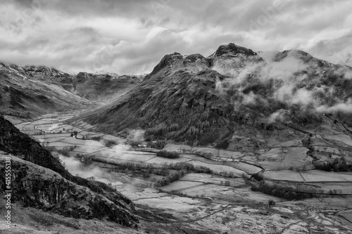 Stunning flying drone black and white landscape image of Langdale pikes and valley in Winter with dramatic low level clouds and mist swirling around